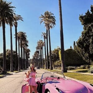 Woman with pink car under palm trees.